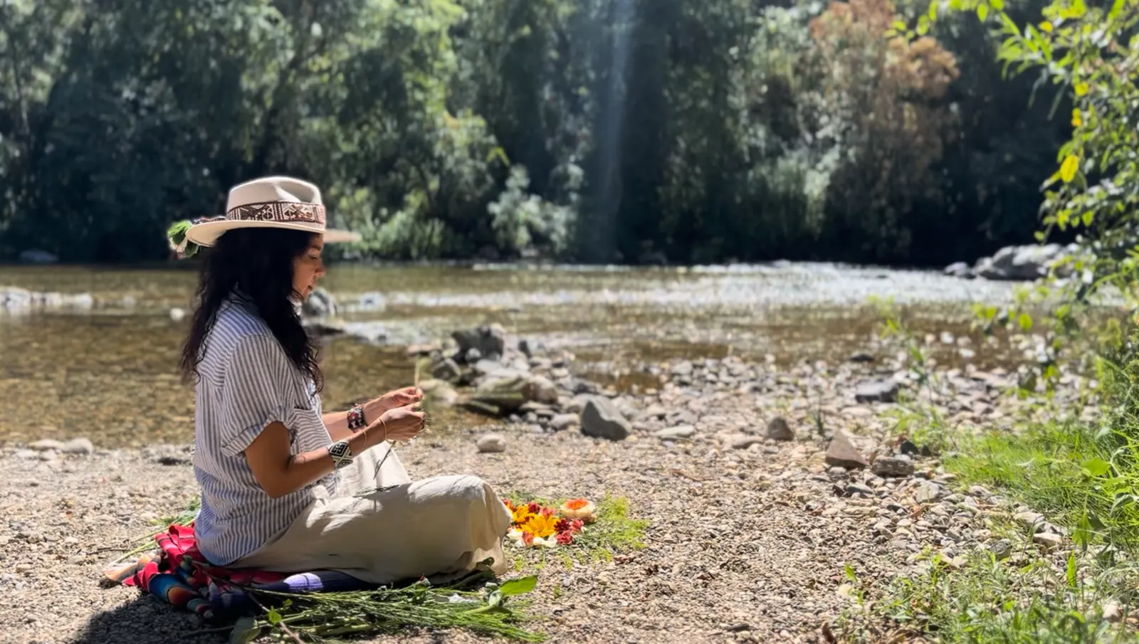 Aimi seated by a river, weaving an offering of flowers and herbs
